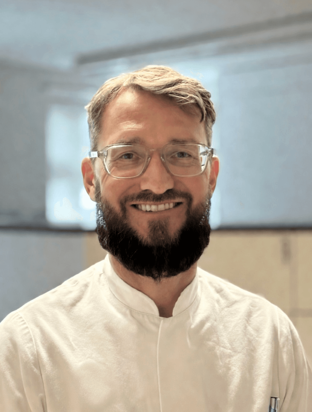 Lächelnder Mann mit Brille und dunklem Bart trägt ein weißes Hemd vor hellem Hintergrund.